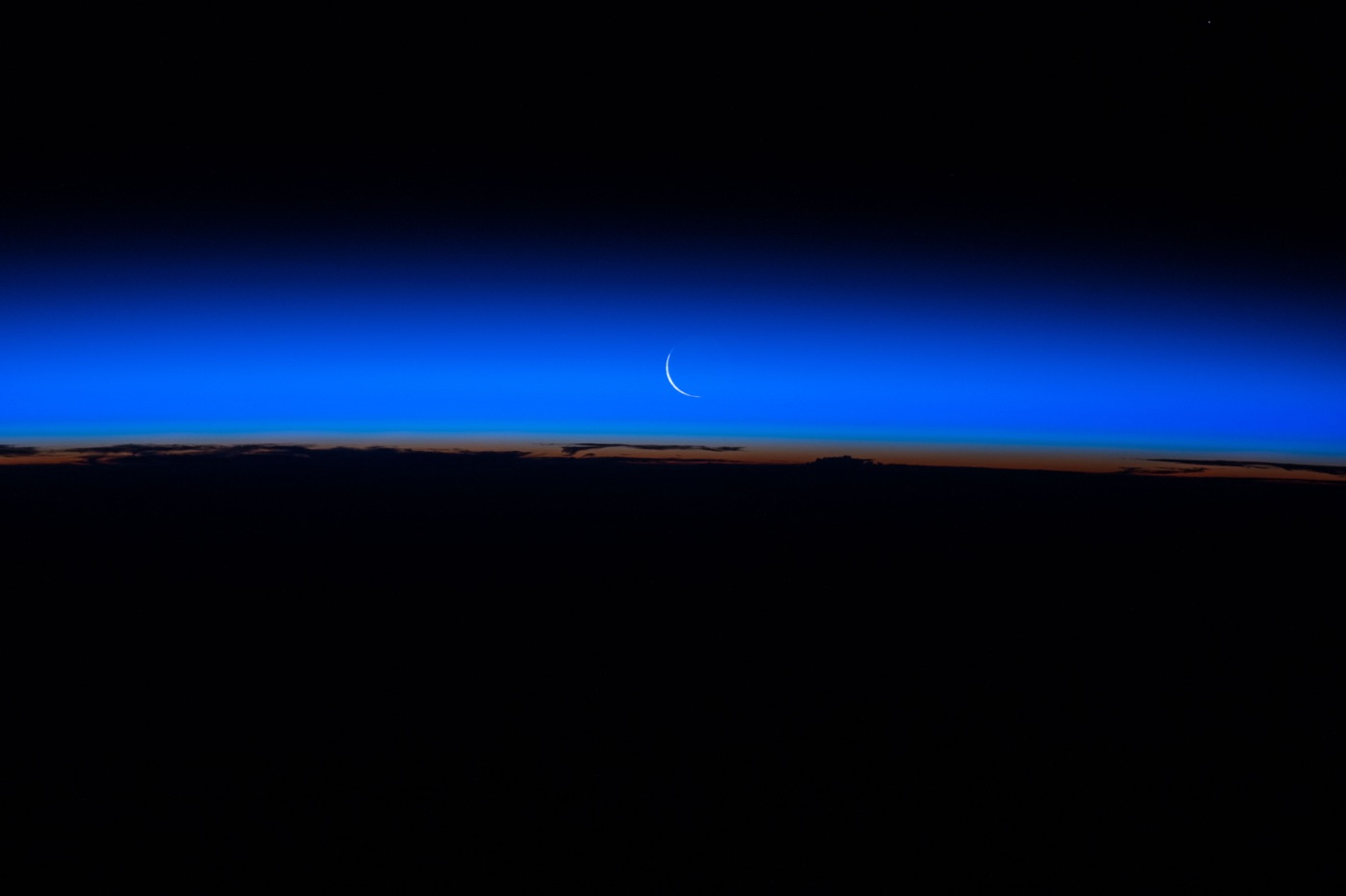 A very thin waxing crescent Moon low in a graded blue twilight band above Earth’s dark limb, with an orange line of sunset below, photographed from the International Space Station.
